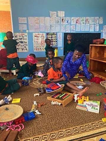 A teacher playing with young learners on the classroom carpet, toys and counting frames around them