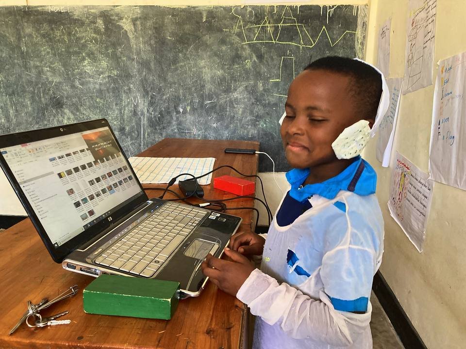 A boy in a white apron and a recycled-material headset working at a laptop beside a hand-built device