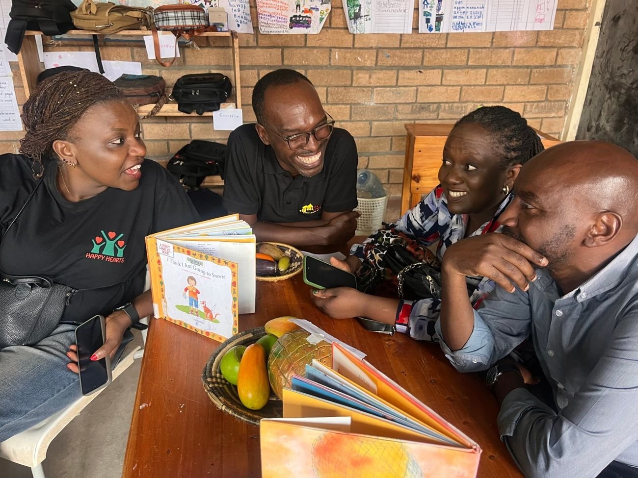 Four colleagues sharing a laugh around a table of picture books and fruit in a classroom