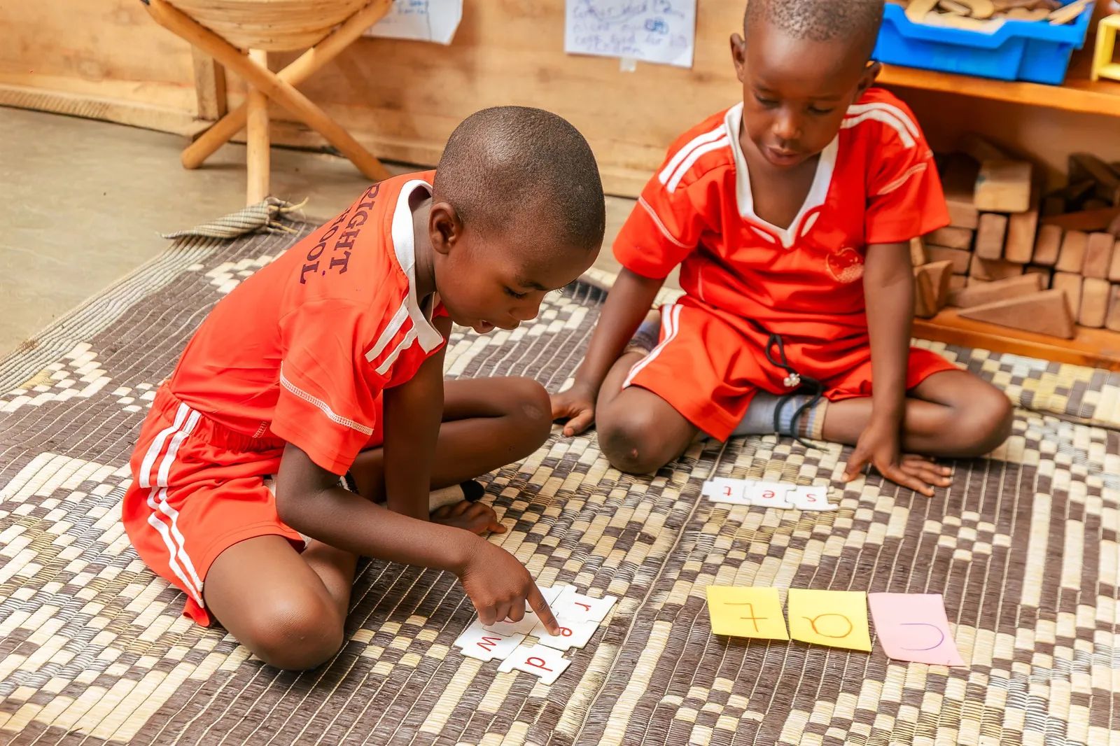 Two boys in red kits building words with letter puzzle pieces on a woven mat