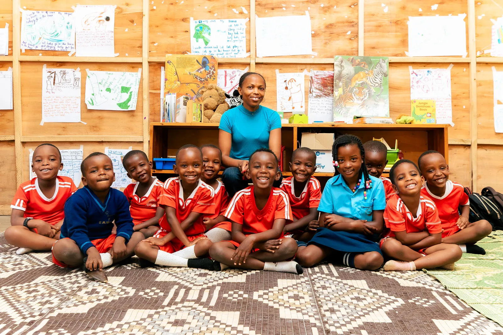 A class of smiling children sitting around their teacher on a patterned mat