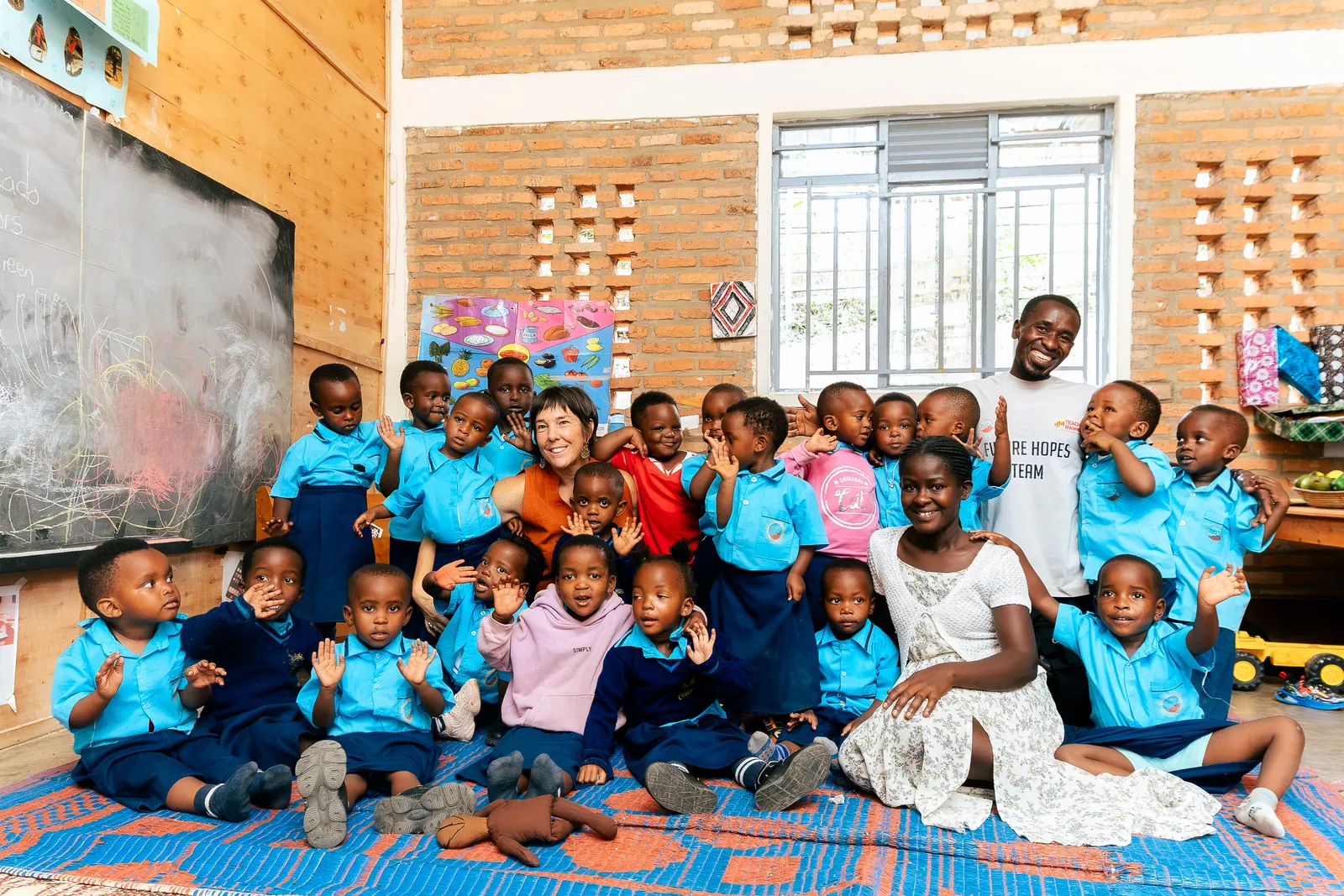 A class of smiling children and visitors sitting together on a colourful mat