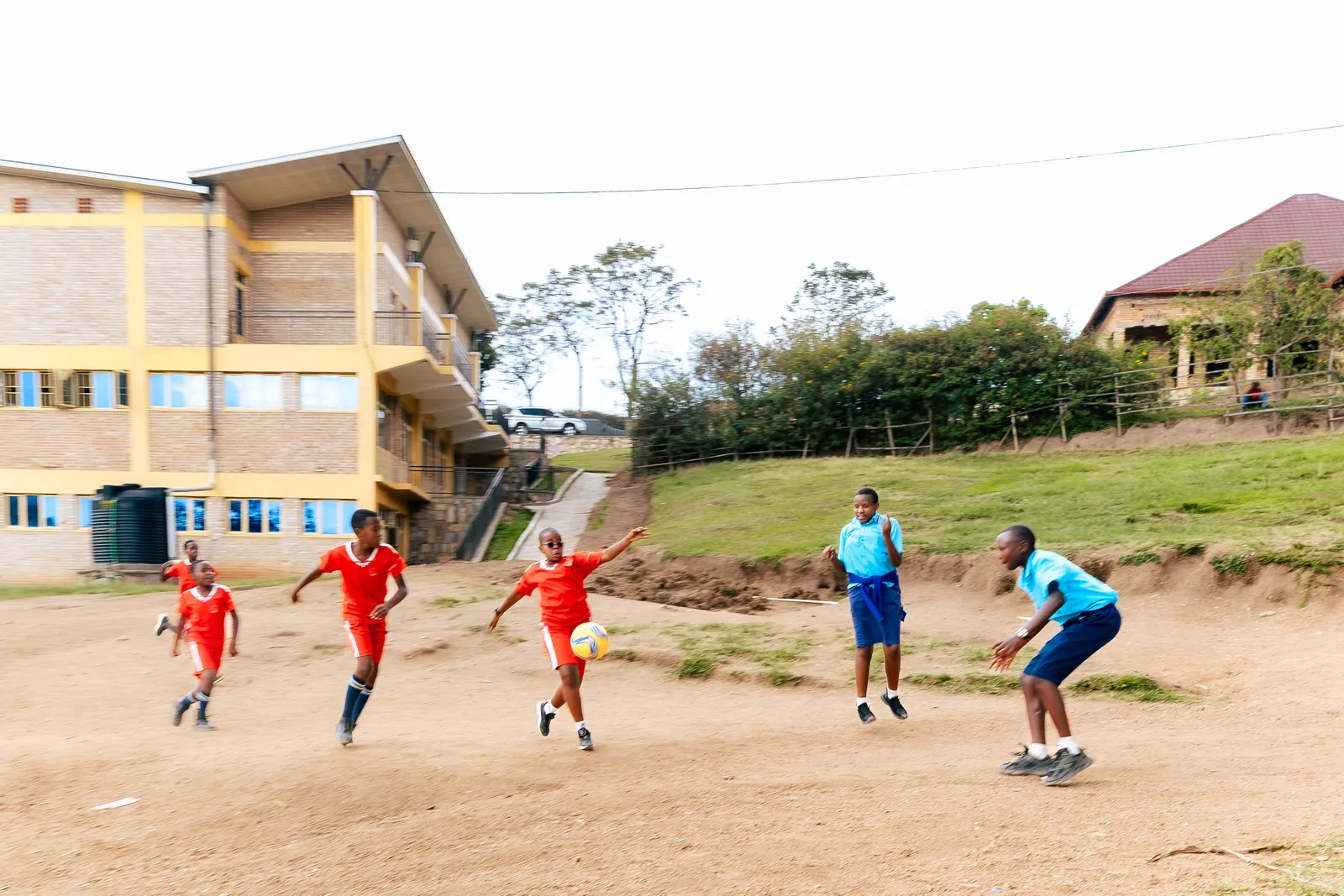 Children in red and blue playing football on the pitch in front of the school