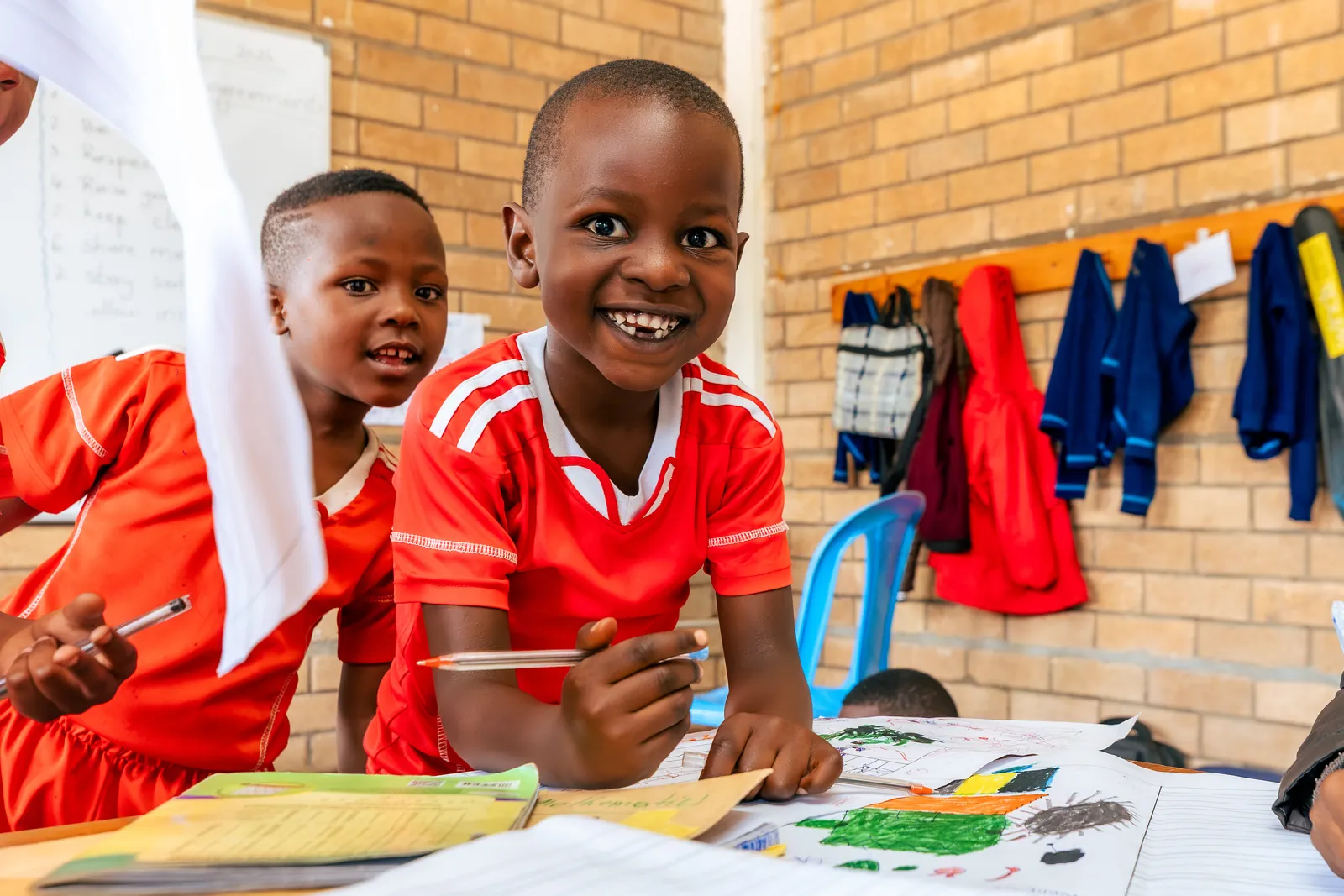 A girl in a red shirt grinning broadly while drawing at a table