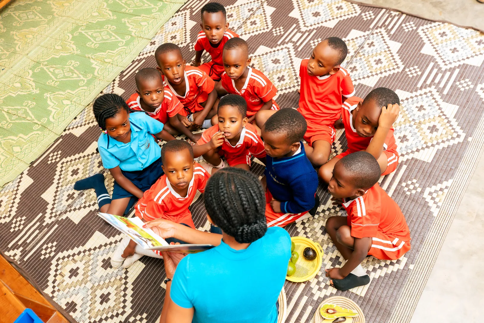 Children in red and blue seated on a patterned rug, looking up at the camera