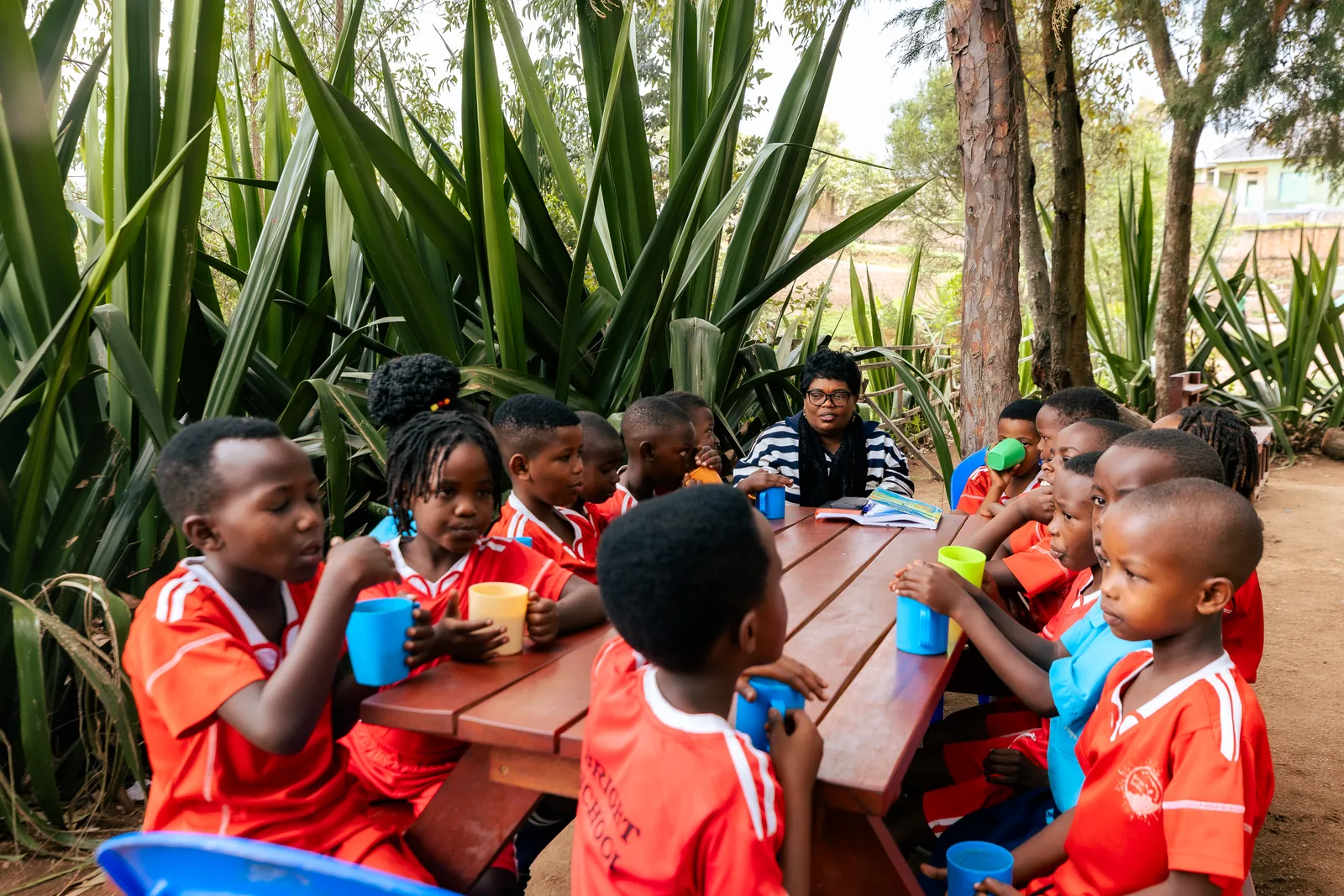 Children and a teacher sitting around a red table among tall green plants