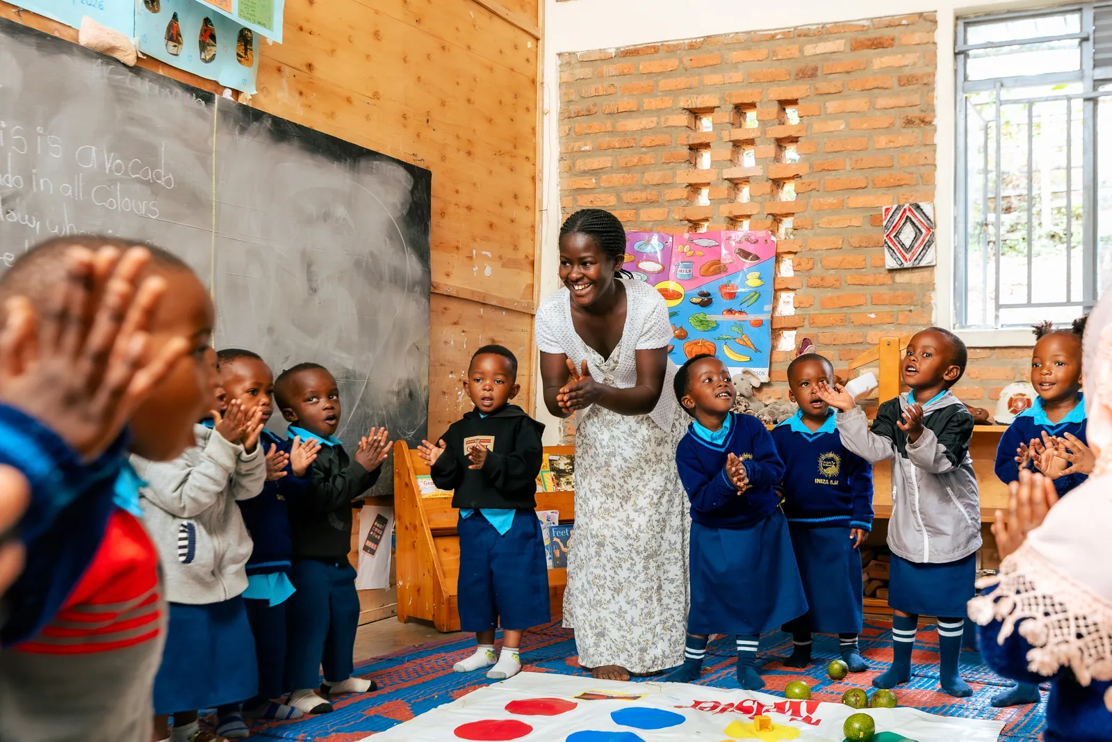 A teacher clapping with a class of children standing around a play mat
