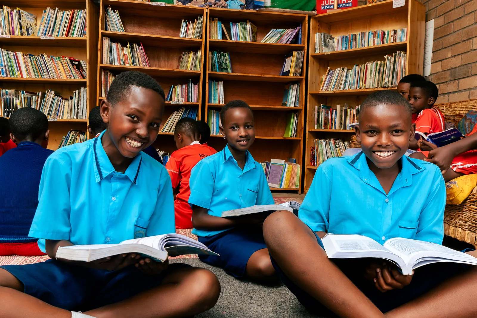 Three smiling children in light blue shirts holding open books in the library