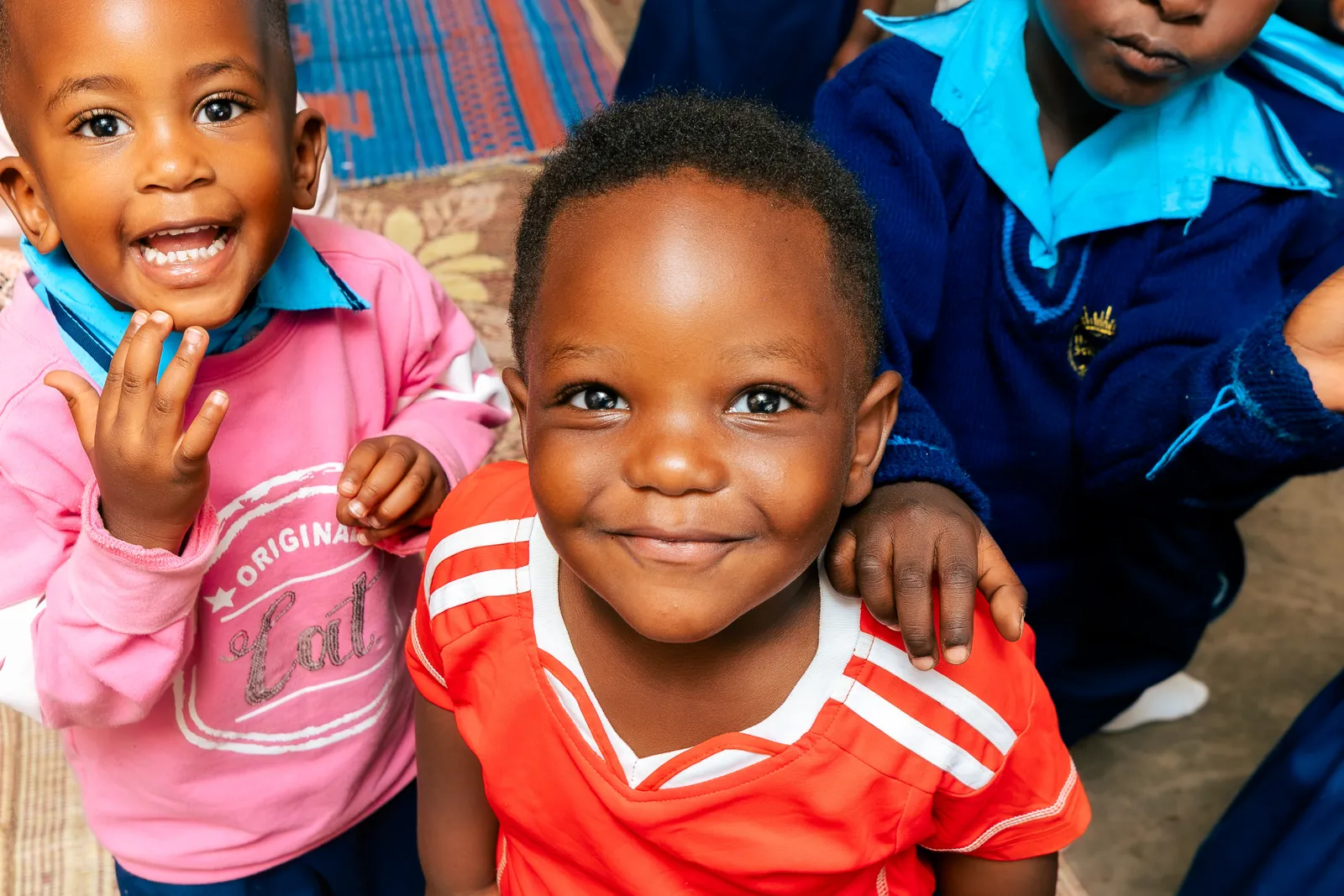 Close-up of a smiling toddler in a red kit with a friend waving behind