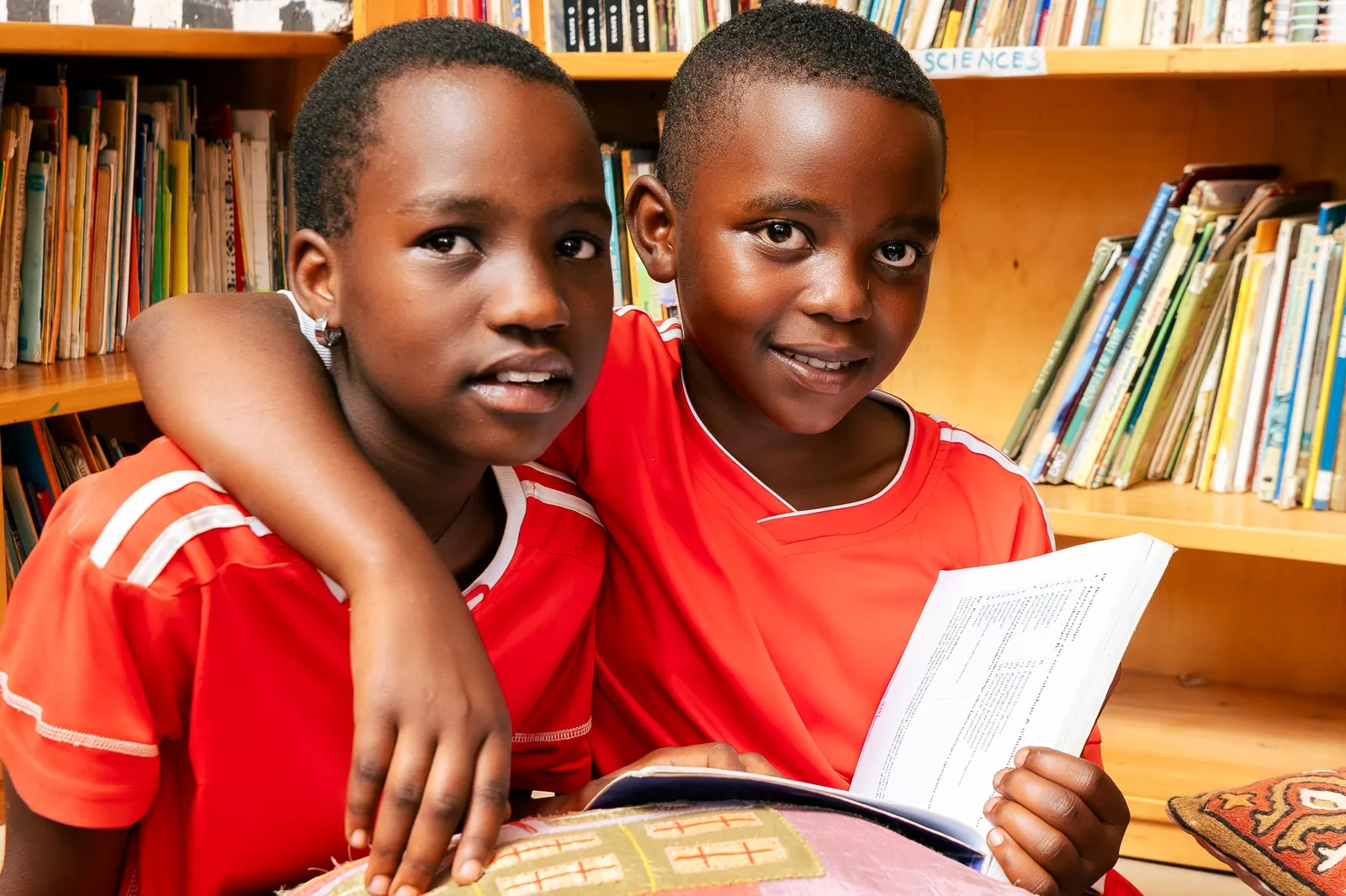 Two children with arms around each other holding a book in front of shelves